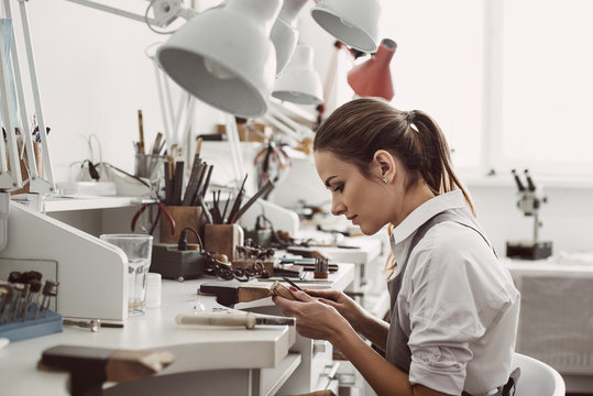 Working all day. Side view of young female jeweler sitting at her jewelry workshop and holding in hands jewelry tools for making accessories