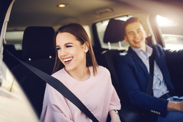 Elegantly dressed smiling Caucasian brunette driving in car and looking through window while her boyfriend driving.