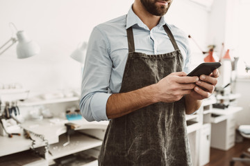 I manage all process online. Close up portrait of young male jeweler in apron holding a smartphone