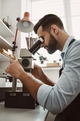Maximum accuracy. Side view of a male jeweler looking at the ring through microscope in a workshop.