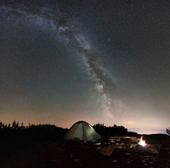 Tourist camping at night on the top of rocky mountain. Glowing tent and bonfire under incredibly beautiful night sky full of stars and Milky way. On background amazing starry sky and mountains