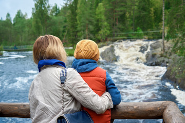 Mother and son are looking towards the wild rough rive form the wooden bridge. They are walking around picturesque nature reserve.