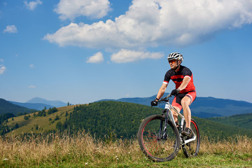 Professional sportsman cyclist in sportswear and helmet cycling a bike in high grass, enjoying beautiful view of Carpathian mountains on sunny summer day. Active lifestyle and extreme sport concept