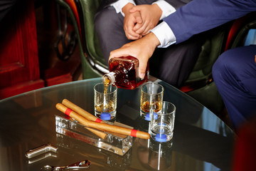 man pours alcohol in three glasses, three cigars lie in an ashtray