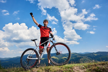 Happy professional sportsman cyclist standing with cross country bicycle on a hill, rasing hand,...