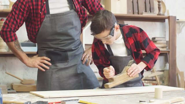 Carpenter watching his little trainee working with jointer at the wooden table with diverse working tools laying on it
