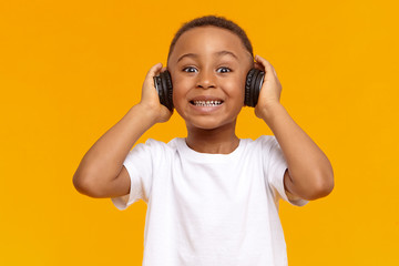 Isolated shot of funny toothy dark skinned little kid in white t-shirt posing at yellow studio wall in wireless headset, listening to music and smiling, having happy pleased facial expression