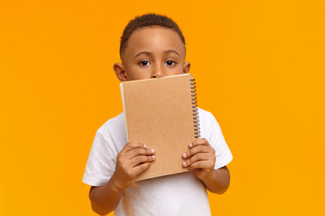 Children, lifestyle, education and learning concept. Portrait of cute handsome African American schoolboy in white t-shirt posing isolated in studio, holding copybook with blank copyspace cover