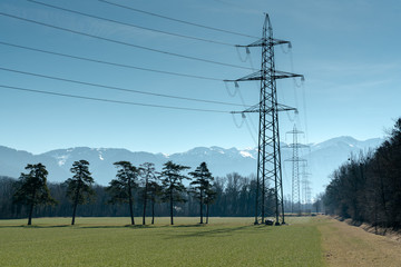 high voltage power lines and tall lattice crosses in rural countryside with snow-capped mountain peaks in the background