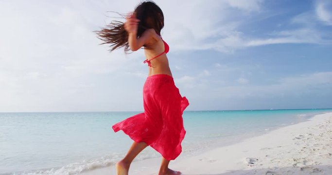 Joy and Happiness. Happy beach woman having fun running dancing on beach - weight loss or travel concept. Joyful girl wearing red bikini and sarong jumping of joy in beachwear. Slow motion shot on RED