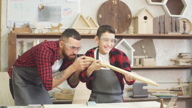 Little male woodworker creating his dream and his Dad is helping him. Close-up of young male carpenter examines with his son smoothness of DIY wooden sword to work with wood in his workshop