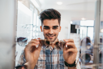Handsome young man choosing eyeglasses frame in optical store.