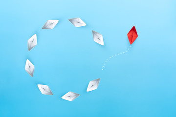 Group of white paper ships float in a circular direction and one red paper ship pointing in...