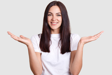 Isolated shot of joyful brunette pretty woman laughing happily when she sees or hears something funny, has medium dark hair, poses on gray studio wall. Concept of happiness and positive emotions.