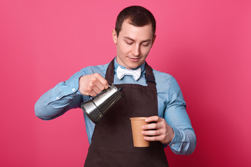 Waist up shot of professional male barista pours coffee from coffeemaker into paper cup, wears shirt and apron, white bowtie poses against pink background, enjoys pleasant aromat of hot beverage