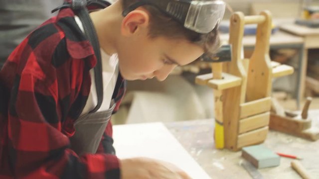 Hardworking little caucasian boy in safety glasses and an apron works with chisel in a carpentry shop being supervised by adult professional woodworker.