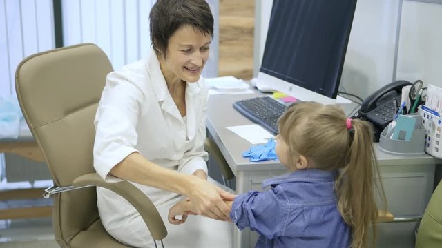 Smiling female children doctor rolling up shirt sleeve of little girl and talking to her while palpating her arm