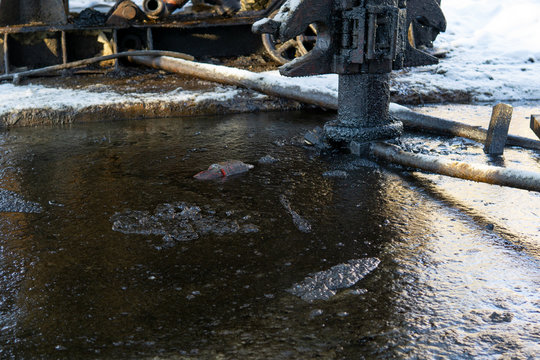 A Close Up Of The Surface Of Black Oil Pollution On Water. Closeup Of An Oil Slick In Water With Fall Colors In The Grass On The Shore