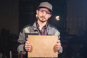 Portrait of a young carpenter in overalls and goggles with a mock up board holding a blank sign in his hands in his home workshop