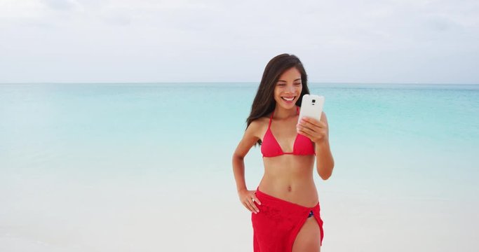 Beach vacation travel woman taking smart phone selfie in bikini having fun sharing on social media. Self portrait photo with smartphone on beach. Happy mixed race Caucasian / Asian Chinese woman.