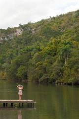 Young blonde woman in summer dress at pier near wild river in jungle