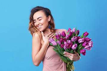 Young happy smiling redhead woman holding bouquet of colorful spring flowers isolated on blue background. Pink tulips, festive bouquet in honor of women's day on March 8 or birthday