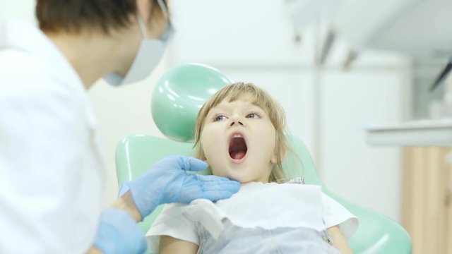 Little girl sitting in dental chair and opening her mouth, female dentist in medical gloves examining her teeth