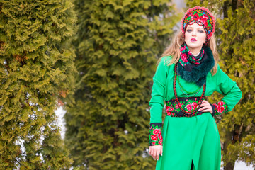 Obraz premium Caucasian Blond Girl in Fashionable Green Dress and Kokoshnik with Flowery Pattern and Beads. Posing Against green Trees in Park Outdoors.
