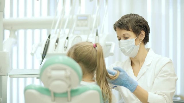Female dentist in mask and uniform helping anonymous girl to sit down on professional chair in clinic