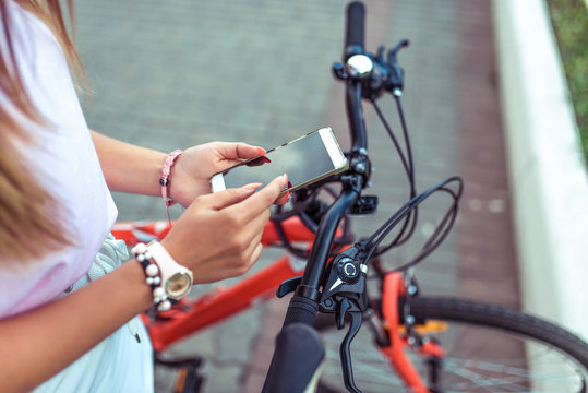 Girl is holding smartphone, searching route through application in Internet map. Against the background of red black bicycle in parking lot. Online in social networks, communication correspondence.