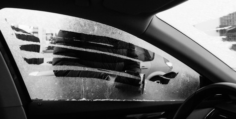 View from inside the car of woman cleaning car driver window from frost using specialized ice frost scraper and rubber squeegee car windscreen in cold winter - black and white.