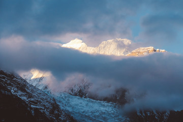 Sunlight wins over clouds above the Himalayan Peaks, Annapurna Circuit Trek, Nepal. Slopes covered with mist. Sharp slopes. Smaller mountain in front, covered in shadow.  Overcast but sunny.