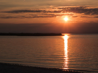 Sunset over the lake. Sun goes down behind the clouds. Sunbeams reflecting in the calm surface of the lake. A small island in the middle of the lake. Neusiedlersee in Austria. Pebbles beach.