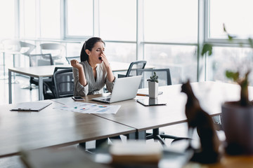 Sleepy young business woman sitting at office desk, working with a laptop and yawning