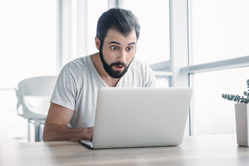 Fototapeta premium Young stressed businessman sitting with laptop with shocked facial expression. Surprised business man looking at laptop, computer worried and amazed with open mouth and big eyes