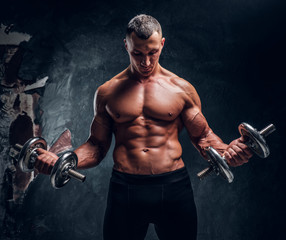 Fototapeta na wymiar Muscular young guy trains with two dumbbells. Studio photo with dark wall background