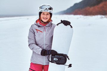 Fototapeta na wymiar Happy Norwegian girl wearing warm snowboard suit, posing with her snowboard on a snowy beach