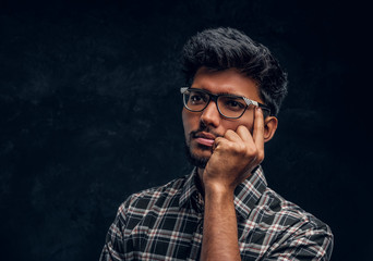 Pensive Indian student wearing eyewear and a plaid shirt. Studio photo against a dark textured wall