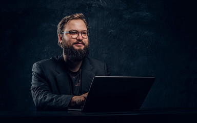 Smiling bearded programmer sitting at the table with laptop in office against a dark wall