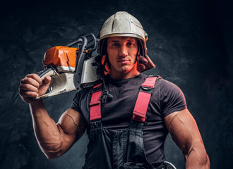 Brutal logger wearing protective clothes and helmet posing with a chainsaw on his shoulder and...