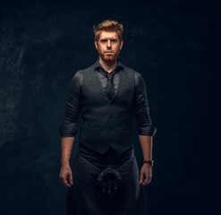 Handsome redhead man dressed in an elegant vest with tie and kilt in studio against a dark textured...