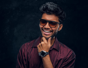 Vogue, fashion, style. Cheerful young Indian guy wearing a stylish shirt and sunglasses posing with hand on chin. Studio photo against a dark textured wall