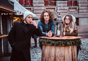 Two guys and girl tourists posing on camera smiling drinking coffee on the market of the city square
