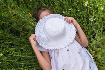 A young girl in a white dress and with a hat in her hands is lying on a green meadow with daisies
