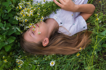 Beautiful girl with blond hair in a white dress lies on a green meadow with a bouquet of daisies