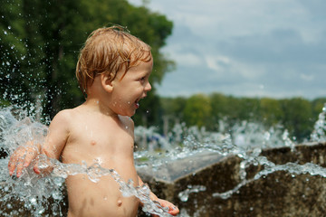 Funny baby boy trying to cauch water stream in fountain. Cute toddler playing in the city fountain