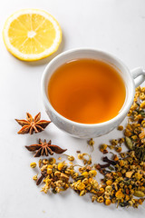 A Cup of green tea with dried chamomile flowers and a slice of cut lemon on a white background....
