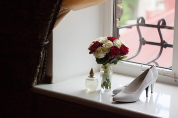 bridal bouquet of red and white roses in a glass vase, shoes and a bottle of perfume on the windowsill