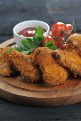 Chicken wings in crispy breading on a wooden board. The composition is complemented with cherry tomatoes, parsley and a cup with ketchup sauce. Close-up. Macro shooting. Vertical frame orientation.