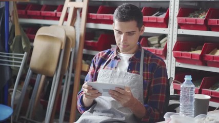 Handsome young man in apron looking at camera and smiling while sitting near shelves in studio and using modern tablet - Powered by Adobe